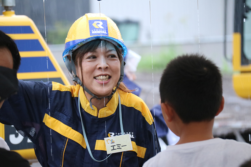 雨ニモ負ケズ、元気ハツラツで子どもを誘導するエミーさん。真剣な眼差しで重機の動きを追うこども。