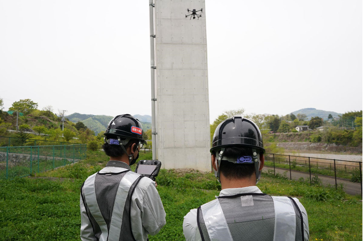 峠下社長（左）。コンクリート橋脚の点検画像をドローンで取得している。機体は市販のDJI社のMatrice300RTKとし、カメラと自動飛行撮影アプリならびに精度管理アプリを自社開発として、点検品質とコストダウンの両立を図っている。自動飛行撮影アプリと精度管理アプリにより現場での作業効率が向上し、2名での運用が可能となった。