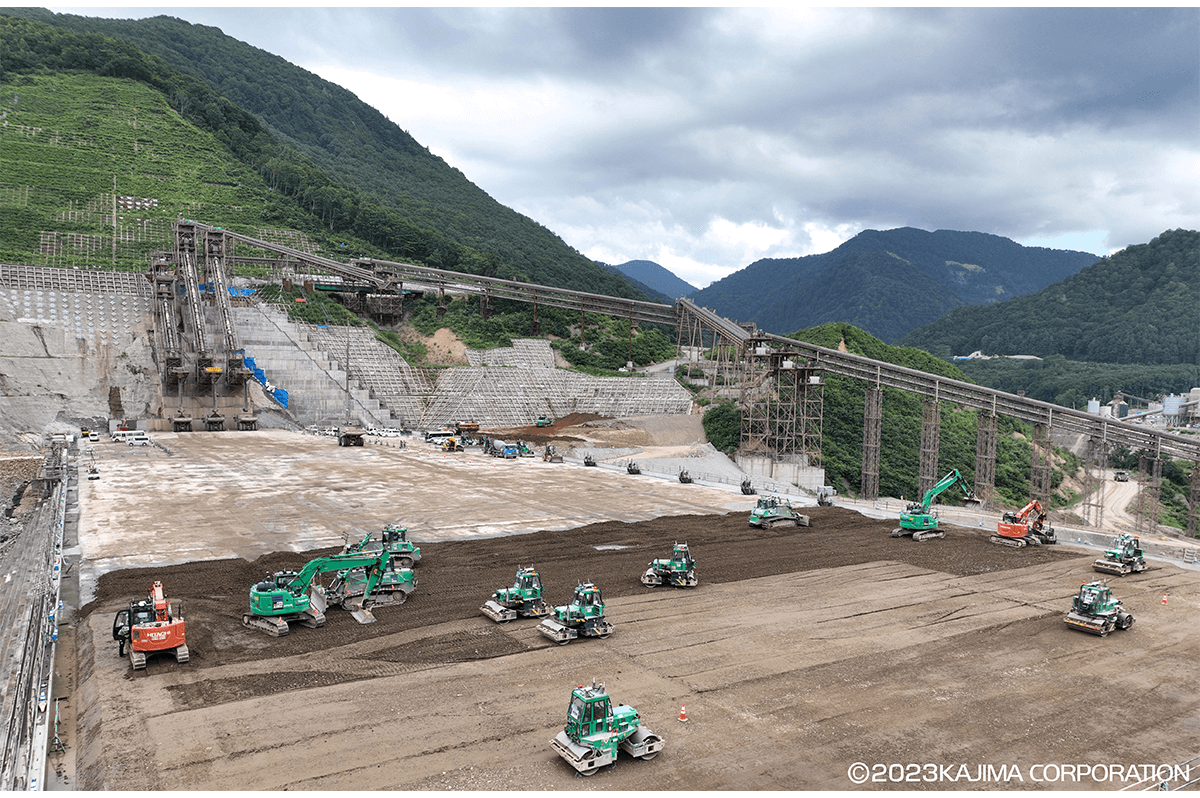 2023年8月、成瀬ダムにおける自動施工の様子。自動重ダンプによるCSG（現地発生材とセメント、水を混合してつくる材料）の運搬、自動ブルドーザによる巻き出し・敷均し、自動振動ブルドーザによる締固めが行われている。なお、一部重機は有人。（鹿島建設写真提供）